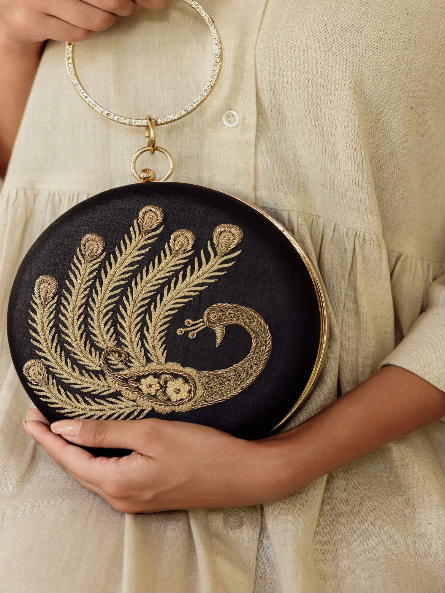 A person in a cream-colored linen dress holds a round black clutch bag with gold peacock embroidery and a gold-tone ring handle.